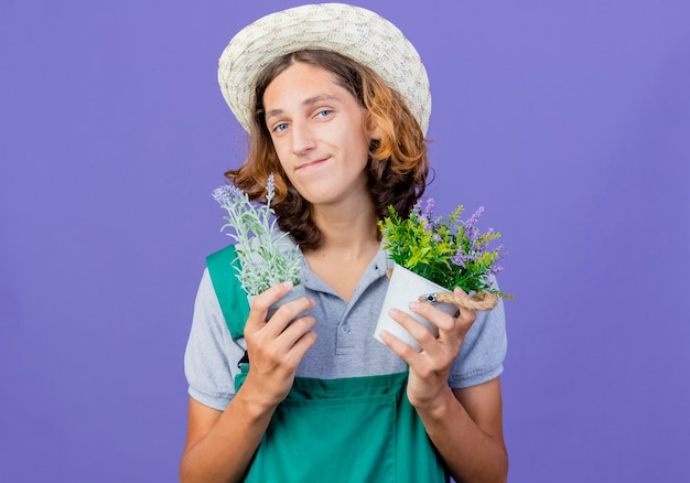 Junger Gärtnermann, der Overall und Hut hält Topfpflanzen hält