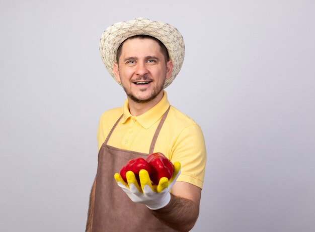 Junger gärtnermann, der overall und hut in arbeitshandschuhen trägt, die rote paprika zeigen, die mit glücklichem gesicht lächeln