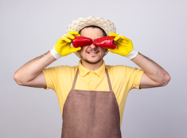 Junger Gärtnermann, der Overall und Hut in Arbeitshandschuhen trägt und rote Paprika über seine lächelnden Augen hält