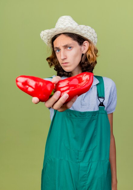 Junger Gärtnermann, der Overall und Hut trägt, die rote Paprika zeigen