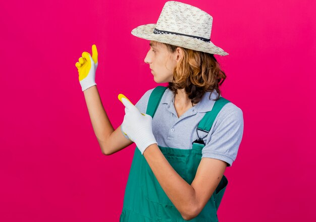 Junger Gärtnermann, der Overall und Hut trägt Gummihandschuhe zeigt