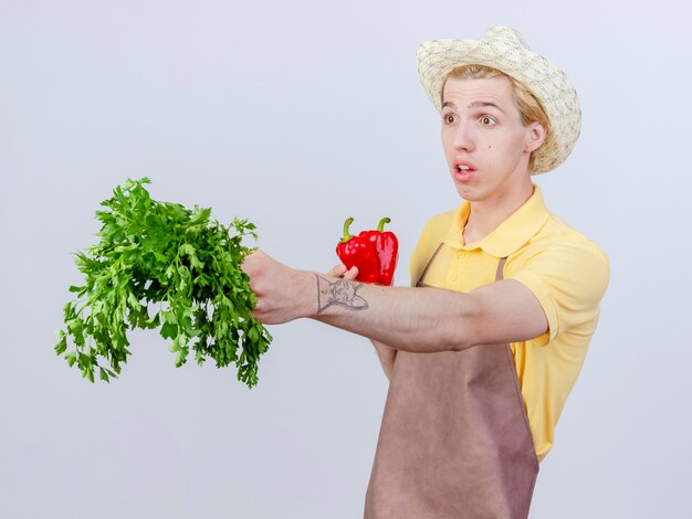 Junger Gärtnermann mit Overall und Hut mit roter Paprika, der frische Kräuter zeigt, die überrascht beiseite schauen