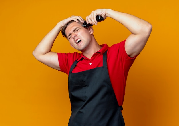 Junger genervter blonder männlicher Friseur in der Uniform hält kopfschneidendes Haar mit Haarschneidemaschine, die auf orange Raum mit Kopienraum isoliert wird