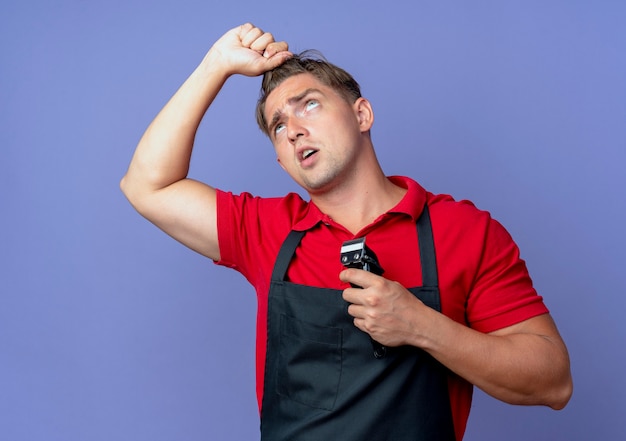 Junger genervter blonder männlicher Friseur in Uniform hält Haare, die nach oben schauen und Haarschneidemaschine isoliert auf violettem Raum mit Kopierraum halten