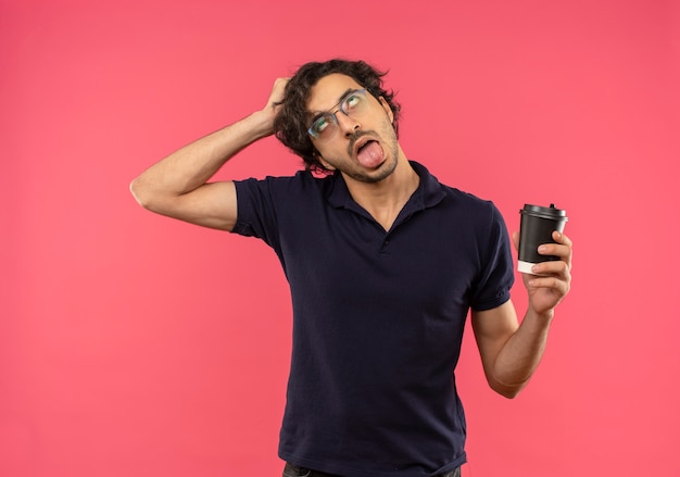 Junger genervter Mann im schwarzen Hemd mit optischer Brille, streckt Zunge heraus und hält Kaffeetasse lokalisiert auf rosa Wand