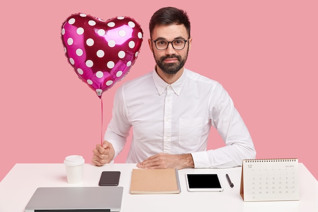 Junger Geschäftsmann, der am Schreibtisch mit Gadgets sitzt und herzförmigen Ballon hält