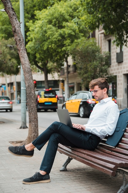Junger Geschäftsmann, der auf Bank über dem Bürgersteig unter Verwendung des Laptops sitzt