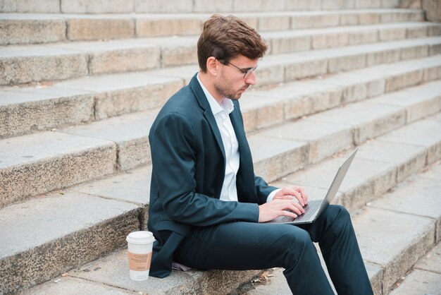 Junger Geschäftsmann, der auf Treppenhaus mit Wegwerfkaffeetasse unter Verwendung des Laptops sitzt