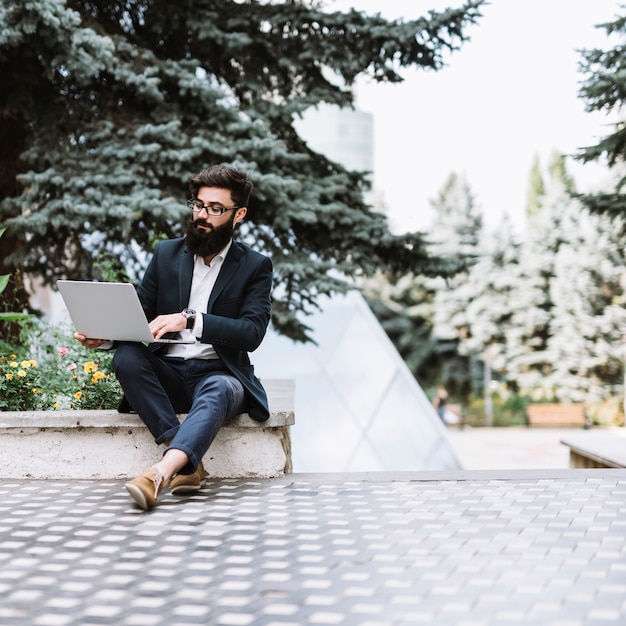 Junger Geschäftsmann, der im Park unter Verwendung des Laptops sitzt