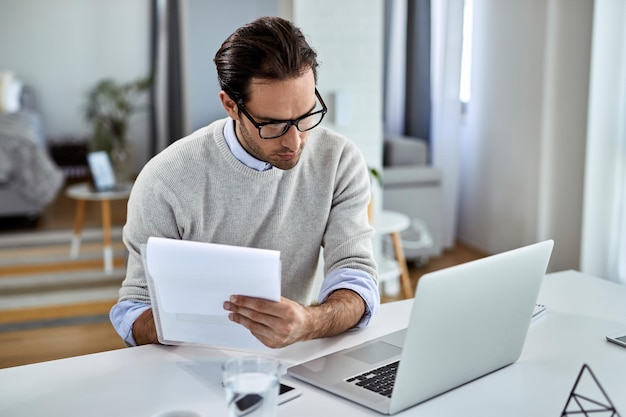 Junger Geschäftsmann geht Papierkram durch und benutzt Laptop, während er zu Hause arbeitet