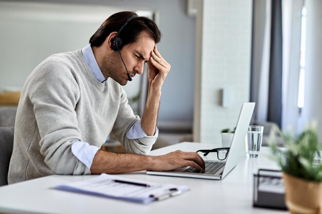 Junger Geschäftsmann hält sich den Kopf vor Schmerzen, während er zu Hause an einem Computer arbeitet