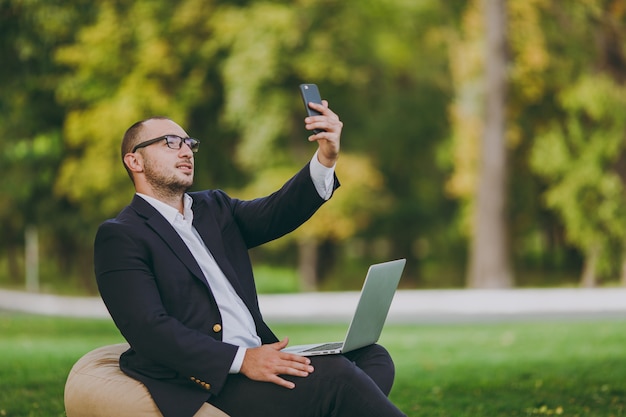 Junger geschäftsmann im weißen hemd, im klassischen anzug, in der brille. mann sitzt auf weichem hocker mit laptop-pc und macht selfie auf dem handy im stadtpark auf grünem rasen im freien. geschäftskonzept für mobiles büro.