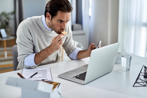 Junger Geschäftsmann isst, während er Handy benutzt und zu Hause am Laptop arbeitet