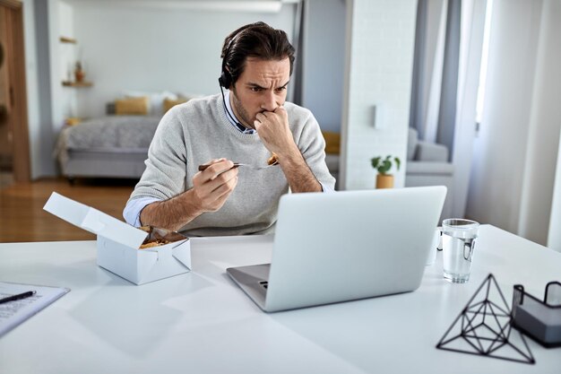 Junger Geschäftsmann isst, während er zu Hause problematische E-Mails auf einem Computer liest