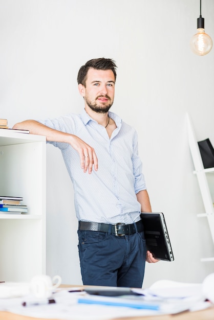 Junger Geschäftsmann mit dem Laptop, der im Büro steht