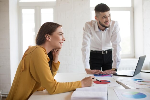 Junger Geschäftsmann und Frau schauen glücklich beiseite, während sie gemeinsam mit Diagrammen im modernen Büro arbeiten