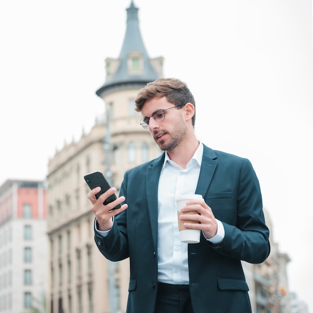 Junger Geschäftsmann unter Verwendung des Smartphone, der in der Hand Kaffeetasse hält