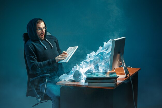 Junger gestresster gutaussehender Geschäftsmann, der am Schreibtisch im modernen Büro arbeitet und Laptop-Bildschirm schreit und über Spam wütend ist