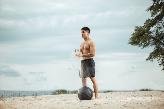 Junger gesunder Mannathlet, der Übung mit Ball am Strand tut