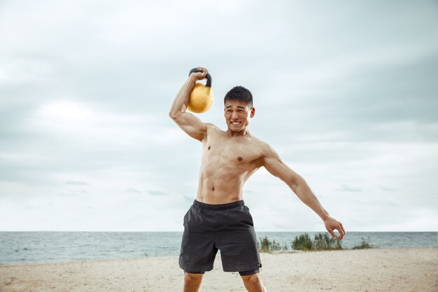 Junger gesunder Mannathlet, der Übung mit dem Gewicht am Strand tut. Hemdloses Training des männlichen Modells des Signels an der Flussseite am sonnigen Tag. Konzept des gesunden Lebensstils, des Sports, der Fitness, des Bodybuildings.