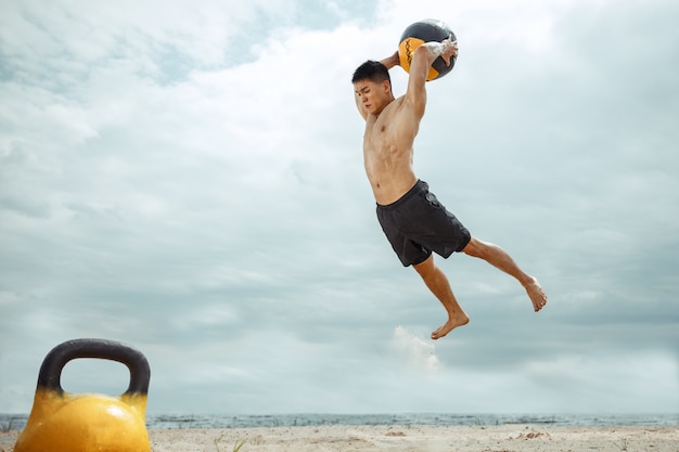 Junger gesunder Mannathlet, der Übung mit dem Gewicht und Ball am Strand tut. Hemdloses Training des männlichen Modells Signle am Flussufer. Konzept des gesunden Lebensstils, des Sports, der Fitness, des Bodybuildings.