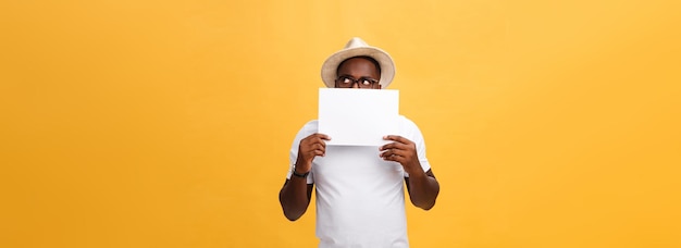 Junger glücklicher Afroamerikaner, der sich hinter einem leeren Papier versteckt, das auf gelbem Hintergrund isoliert ist