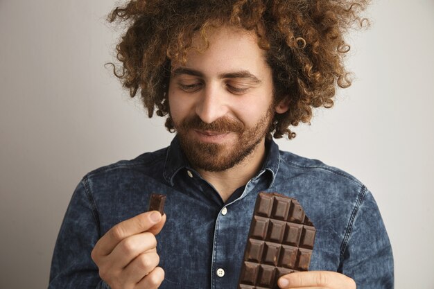 Junger glücklicher bärtiger Mann mit gesunder Haut und lockigem Haar erfreut mit Geschmack der frisch gebackenen Bio-Schokoladentafel
