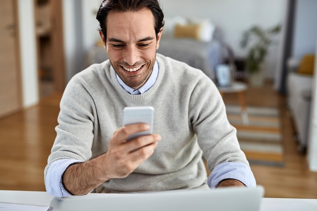 Junger glücklicher Geschäftsmann, der zu Hause arbeitet und SMS auf dem Handy sendet