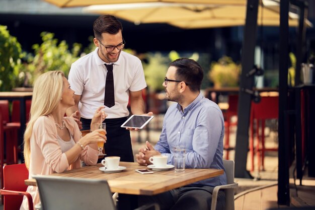 Junger glücklicher Kellner, der mit Gästen in einem Café im Freien kommuniziert