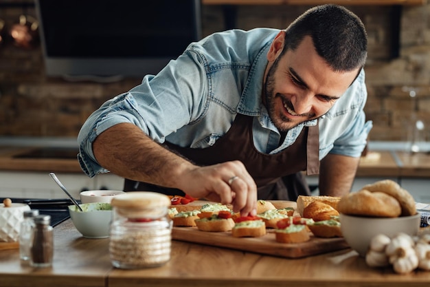 Junger glücklicher Koch bereitet Bruschetta mit Avocadosauce und Kirschtomate in der Küche zu