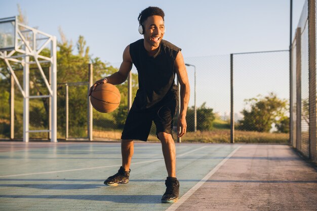 Junger glücklicher lächelnder schwarzer Mann, der Sport macht, Basketball bei Sonnenaufgang spielt, Musik auf Kopfhörern, aktivem Lebensstil, Sommermorgen hört