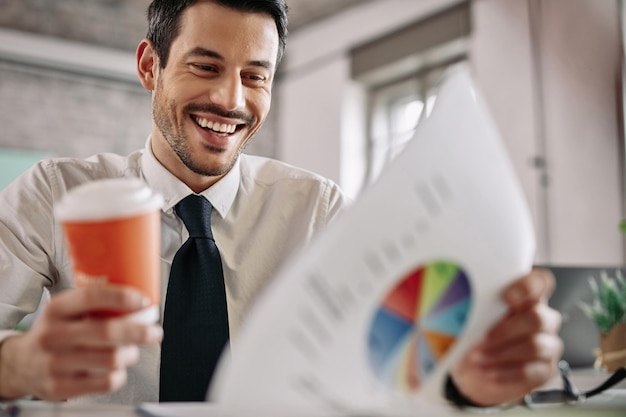 Junger glücklicher Manager, der den Geschäftsfortschritt untersucht, während er Diagramme liest und Kaffee im Büro trinkt