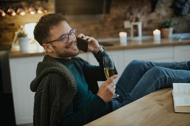 Junger glücklicher Mann, der am Telefon spricht, während er zu Hause Champagner trinkt