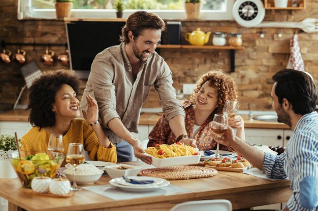 Junger glücklicher Mann, der Essen am Esstisch serviert, während er mit Freunden zu Hause zu Mittag isst.