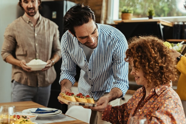 Junger glücklicher Mann, der seiner Freundin Essen serviert, während er mit Freunden zu Hause zu Mittag isst.