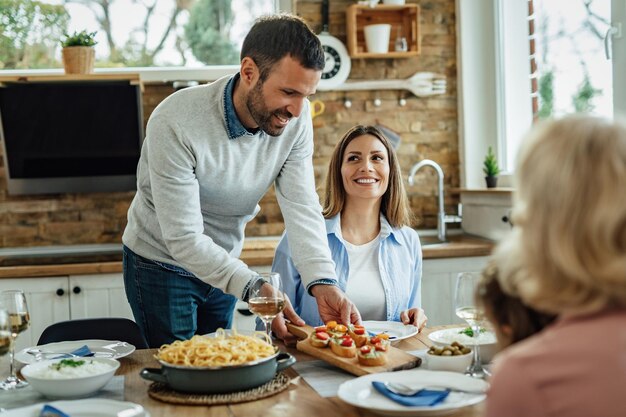 Junger glücklicher Mann serviert Essen und genießt das Mittagessen mit der Familie im Speisesaal.