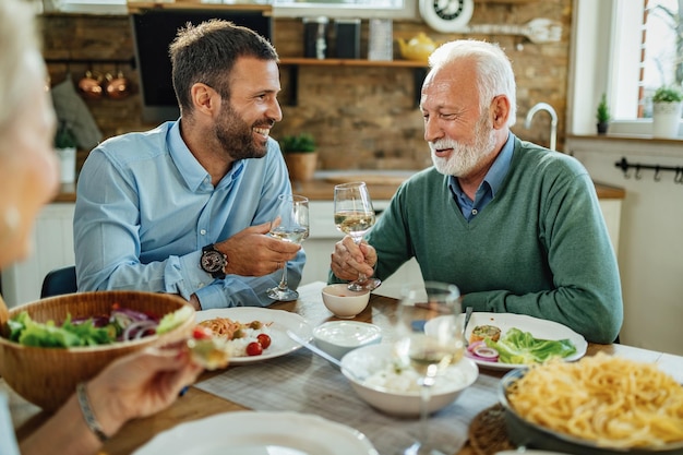 Junger glücklicher Mann und sein reifer Vater stoßen mit Wein an, während sie im Speisesaal zu Mittag essen