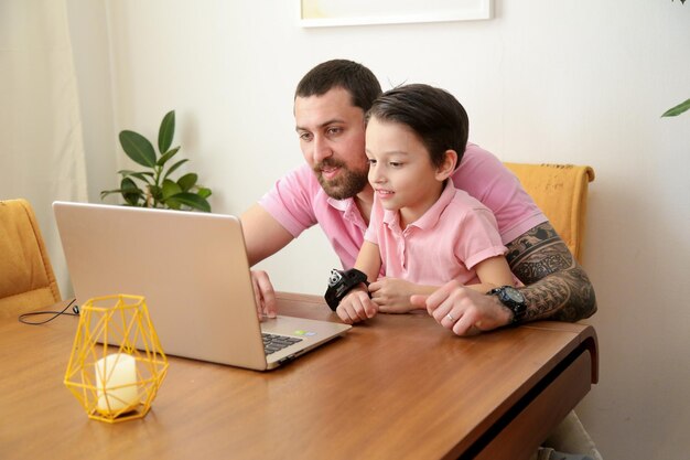 Junger glücklicher vater im rosa poloshirt, der am laptop arbeitet, während er mit seinem kleinen sohn am tisch sitzt glückliche familie, die zu hause arbeitet