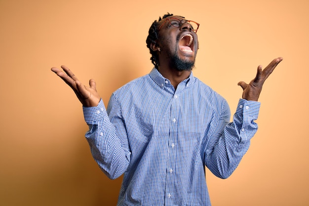Kostenloses Foto junger gutaussehender afroamerikanischer mann mit hemd und brille vor gelbem hintergrund feiert wütend und erfolgsverrückt mit erhobenen armen und geschlossenen augen und schreit aufgeregt nach dem gewinnerkonzept