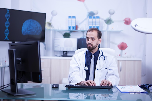 Junger gutaussehender Arzt, der in seinem Kabinett arbeitet und tippt. Doktor, der Tastatur verwendet, um Dokumente auf seinem Computer zu schreiben.