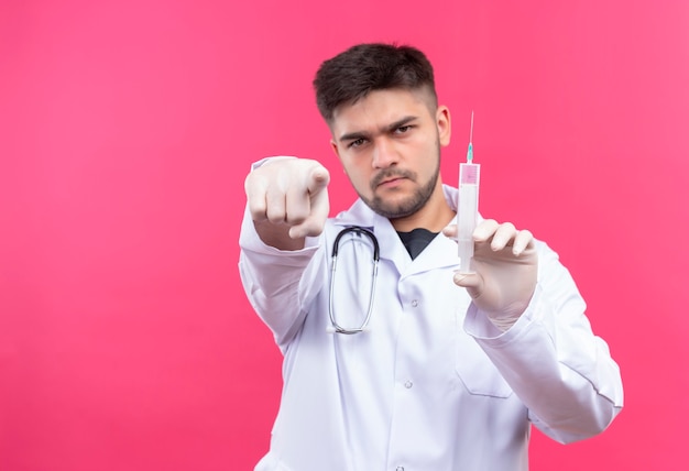 Junger gutaussehender Arzt, der weiße medizinische Handschuhe und Stethoskop des weißen medizinischen Kleides trägt und die Injektion zeigt und hält, die über rosa Wand steht