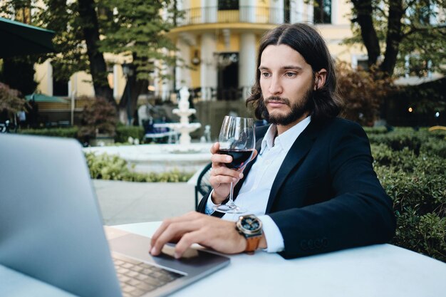 Junger, gutaussehender, bärtiger geschäftsmann mit einem glas wein, der nachdenklich am laptop im restaurant im freien arbeitet