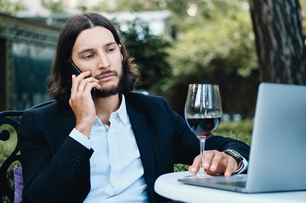 Junger gutaussehender bärtiger Geschäftsmann mit einem Glas Wein, der nachdenklich auf dem Handy spricht, während er mit Laptop im Restaurant im Freien arbeitet