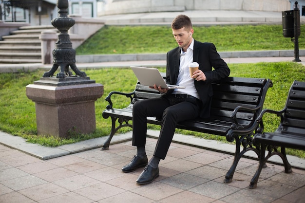 Junger gutaussehender Geschäftsmann im klassischen schwarzen Anzug und weißem Hemd mit drahtlosen Kopfhörern und einer Tasse Kaffee zum Mitnehmen, der nachdenklich am Laptop arbeitet, während er auf einer Bank im Freien sitzt