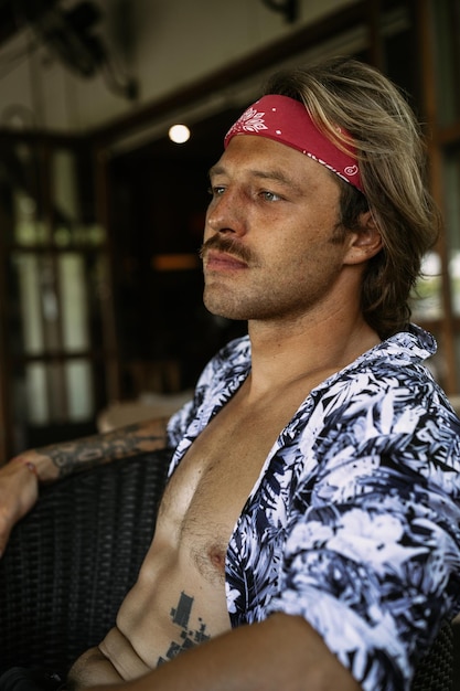 Junger gutaussehender Hippie-Mann in einem roten Halstuch mit nacktem Oberkörper in einem Café in Bali. Glücklicher Mann lächelt