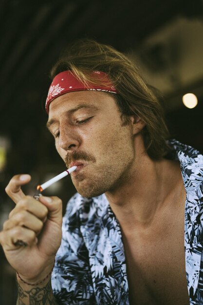 Junger gutaussehender Hippie-Mann in einem roten Kopftuch mit nacktem Oberkörper in einem Café in Bali raucht eine Zigarette