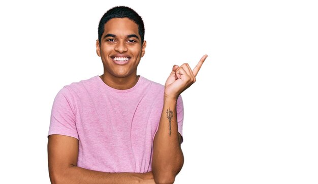 Junger gutaussehender hispanischer mann, der lässiges rosa t-shirt trägt, das glücklich lächelt und mit hand und finger zur seite zeigt