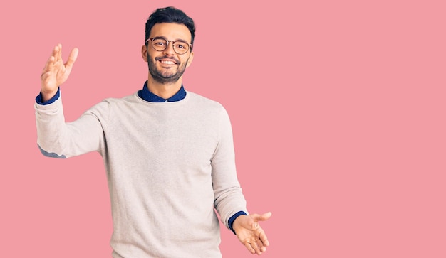 Junger gutaussehender hispanischer Mann in eleganter Kleidung und Brille, der mit offenen Armen lächelnd in die Kamera blickt, um fröhlichen Ausdruck zu umarmen und das Glück zu umarmen