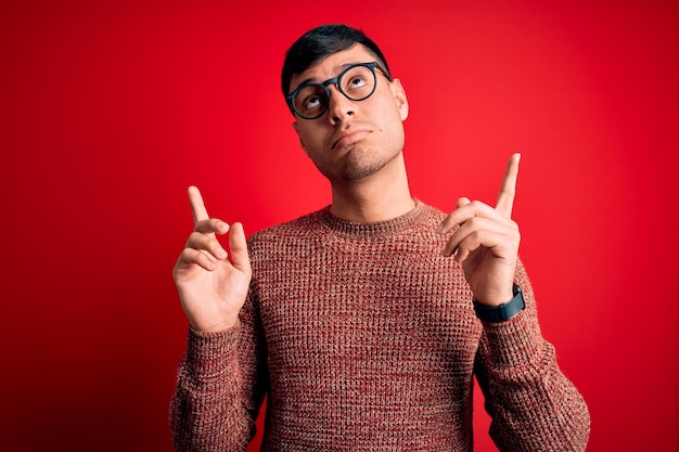 Kostenloses Foto junger gutaussehender hispanischer mann mit nerd-brille vor rotem hintergrund, der nach oben zeigt, traurig und verärgert aussieht und mit unglücklichen und deprimierten fingern die richtung anzeigt