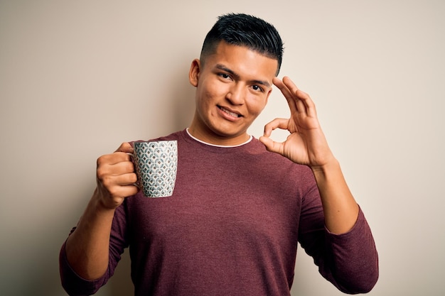 Junger gutaussehender lateinamerikanischer Mann, der vor isoliertem weißem Hintergrund eine Tasse Kaffee trinkt und mit den Fingern ein „OK“-Zeichen macht, ein ausgezeichnetes Symbol